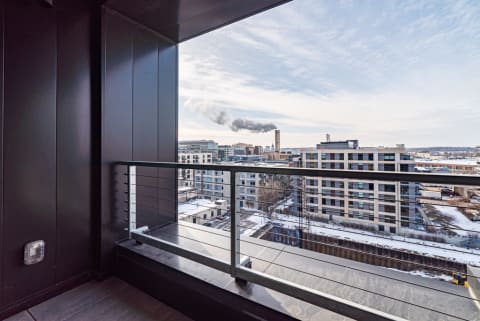 A modern balcony with a view of snowy urban buildings and a smokestack emitting smoke against a cloudy sky.
