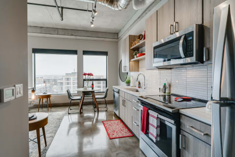 Modern kitchen with light wood cabinetry and stainless steel appliances, featuring a round dining table and urban view through large windows.