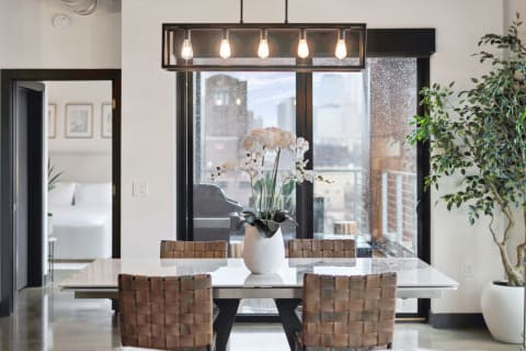 A contemporary dining area with a white table, woven brown chairs, and a large window showing a rainy city skyline.