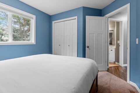 Cozy bedroom with blue walls and white bedding, featuring a window and adjoining bathroom entrance.