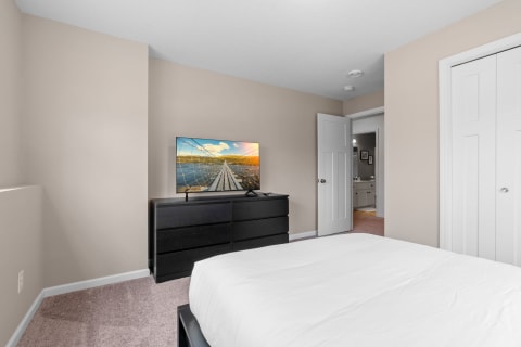 A bedroom featuring a flat-screen TV on a dresser, a made bed, and two doors, with beige walls and soft carpet.