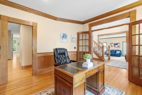 An office with a wooden desk, black chair, and a view of a connected living space with a blue couch.