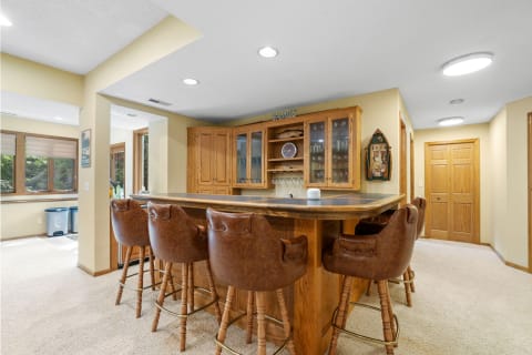 A basement bar area featuring a wooden counter, high-backed chairs, and glass cabinets.