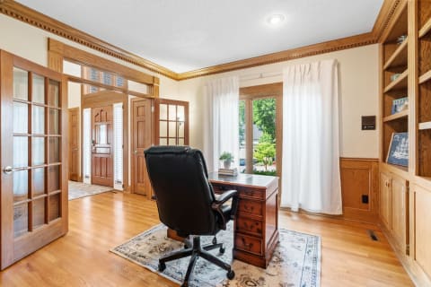 A well-furnished home office featuring a wooden desk, a black chair, and large windows with white curtains.