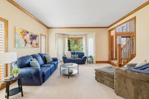 A spacious living room with blue sofas, a round coffee table, and large windows overlooking greenery.