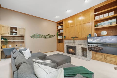 Spacious living room with a gray sectional couch, brick fireplace, and wooden shelves.
