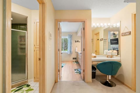 A bathroom hallway featuring a glass shower, vanity with a teal chair, and bright lighting.