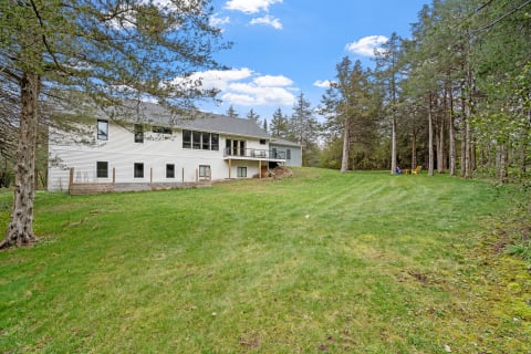A spacious green lawn with a white house, Adirondack chairs, and tall trees under a blue sky.