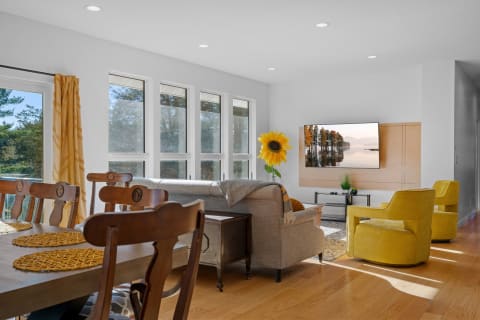 A well-lit living room featuring a gray couch, yellow chairs, and a dining table, adorned with a sunflower and modern decor.