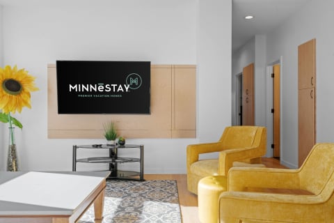Modern living room with yellow chairs, sunflower, and MINNēSTAY logo on a TV.