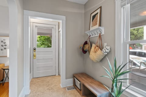 A bright entryway featuring a white door, wooden bench, wall hooks with bags and hats, and a potted plant.