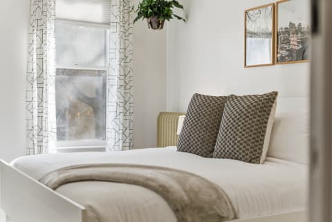 A cozy bedroom featuring white linens, textured gray pillows, and geometric curtains, illuminated by natural light.
