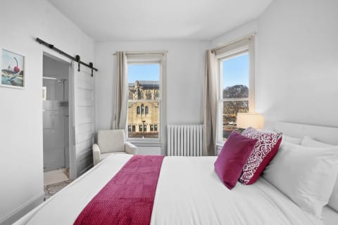 A cozy bedroom with white linens, vibrant pillows, and historic views through the windows.