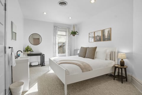 A well-lit bedroom with a white bed, geometric pillows, a round mirror, and nature-inspired decor.