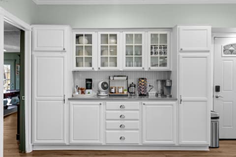 A bright kitchen featuring white cabinets, glass doors with dishware, and a countertop displaying kitchen appliances.
