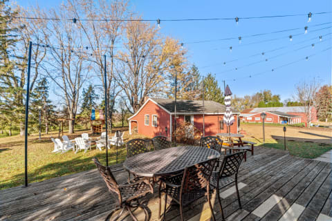 An outdoor dining space featuring a round metal table with chairs, string lights above, and a red house in the background.