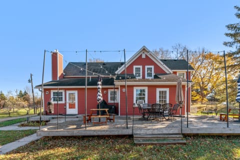 Back view of a red house with a wooden deck, outdoor dining area, and autumn trees in the background.