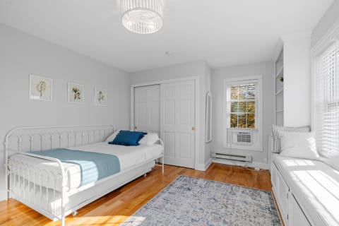 A light gray bedroom with a white metal bed, colorful pillows, and a window seat.