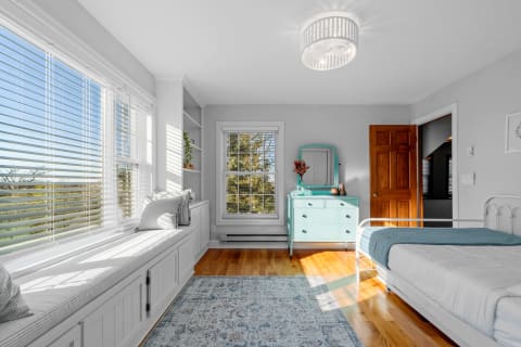 A bright bedroom with a window seat, blue dresser, and wooden flooring.