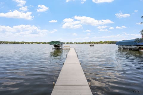 A long dock leading into calm lake water with boats under a green canopy and a blue sky with clouds.