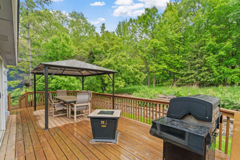 Outdoor deck with a gazebo, dining area, grill, and greenery in the background.