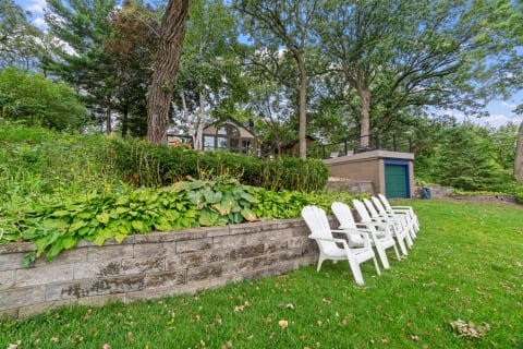 Outdoor setting featuring white Adirondack chairs beside a stone wall and lush greenery.