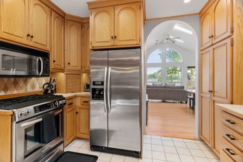 An oak cabinet kitchen with stainless steel appliances, looking into a bright living area through an archway.