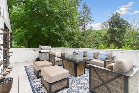Cozy outdoor patio featuring a glass-top table, plush seating, and grill surrounded by trees.