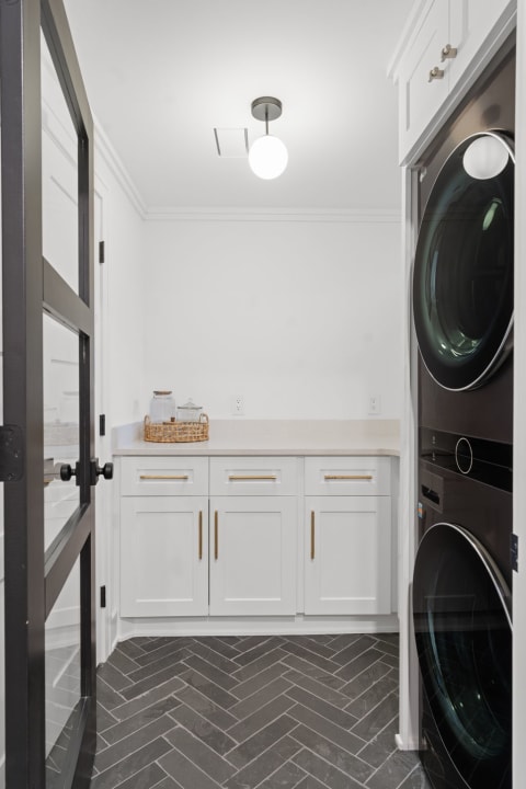 A modern laundry room featuring a black washer and dryer, white cabinets with gold handles, and dark gray herringbone flooring.