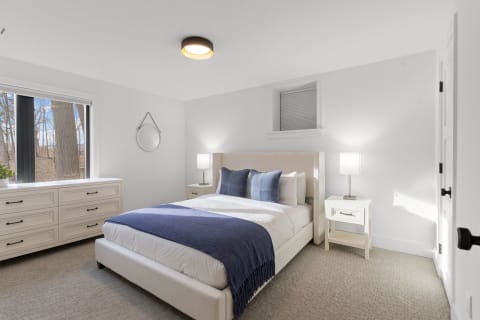 A modern bedroom featuring a white bed with blue accents, nightstands, and a dresser with large windows.