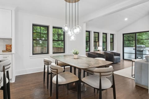 A modern dining area featuring a marble table, plush chairs, and large windows with a view of greenery.