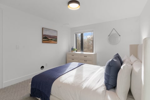 A minimalist bedroom featuring a white bed with a navy throw, light wood dresser, and large window.