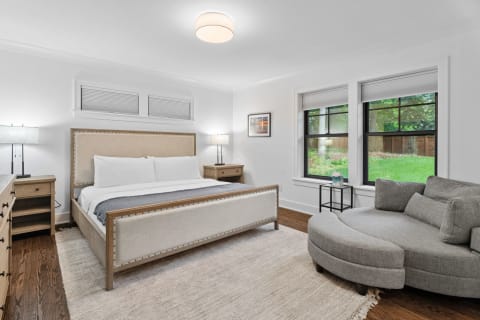 Modern bedroom with an upholstered bed, gray chaise lounge, and large windows overlooking greenery.