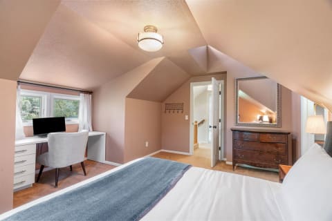 A cozy attic bedroom featuring a workspace, a bed with a gray throw blanket, and a vintage dresser under a slanted ceiling.