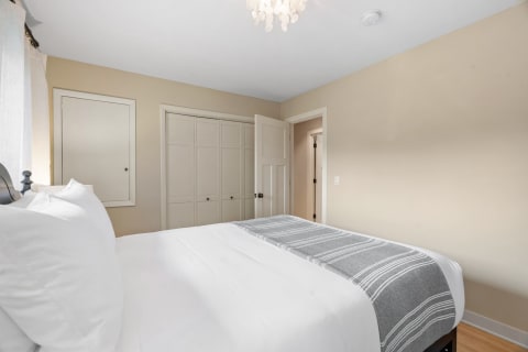A cozy bedroom featuring a bed with white sheets and a grey striped blanket, soft lighting, and warm beige walls.