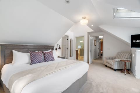 A cozy bedroom with a sloped ceiling, featuring a bed, armchair, TV, and a glimpse of a bathroom.