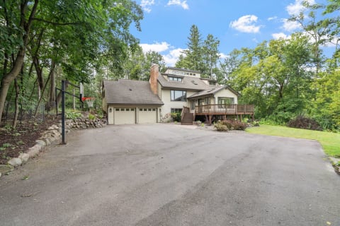 Multi-level suburban house with a deck in a green wooded area and a basketball hoop.