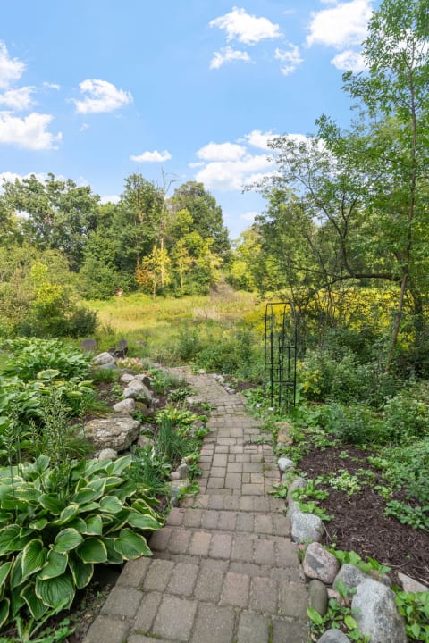 Brick path in a green garden leading to a meadow under a blue sky with clouds.
