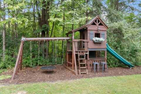 Wooden playhouse with a slide and swing set in a green backyard.