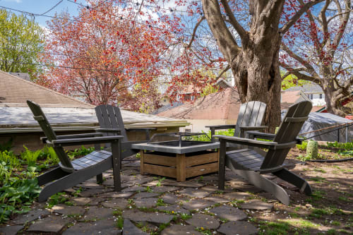 A quaint outdoor seating area with Adirondack chairs and a fire pit surrounded by greenery and spring foliage.