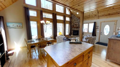 A cozy cabin interior with dining and living areas, under large windows overlooking nature.