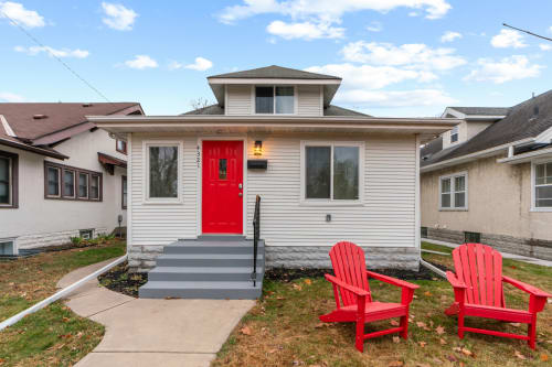 A charming house with a vibrant red door and welcoming Adirondack chairs.