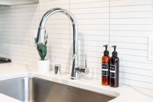 A modern kitchen sink area featuring a sleek faucet and labeled lotion and hand soap bottles.