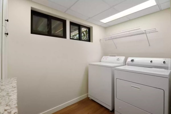 A laundry room featuring a washing machine and dryer beside a wire shelf, with natural light from windows.