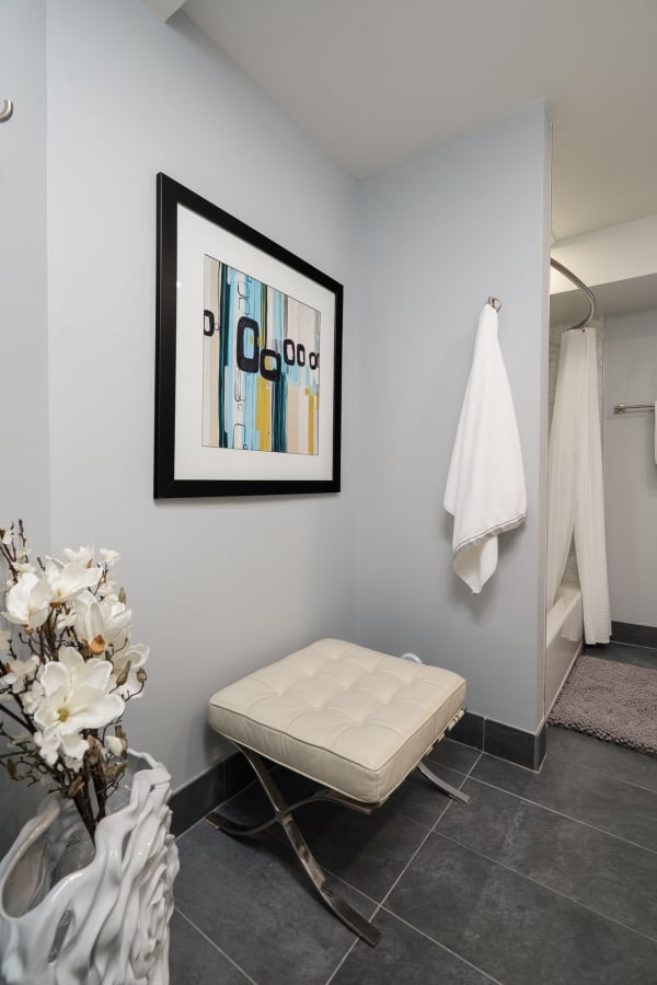 Modern bathroom corner with a white tufted stool, abstract artwork, and a vase of flowers.