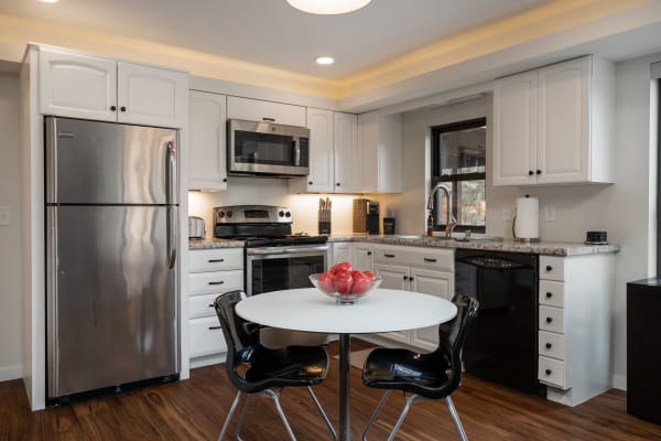 A modern kitchen with white cabinets, stainless steel appliances, and a round table with a bowl of red apples.