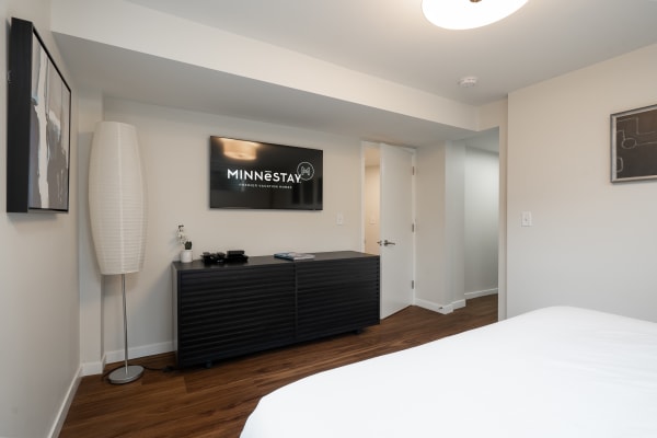 Interior view of a modern vacation rental room with minimalist decor, including a bed, lamp, entertainment unit, and abstract art.
