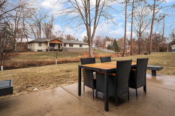 Outdoor dining area with a wicker table and chairs, overlooking a house and trees in a peaceful setting.