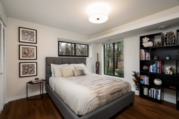 A contemporary bedroom with a grey bed, abstract art on the walls, and a black bookshelf filled with books.