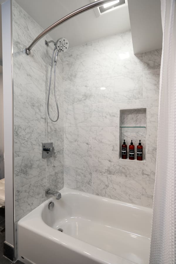 A contemporary shower with marble tiles and a white tub, showcasing bath products on a shelf.